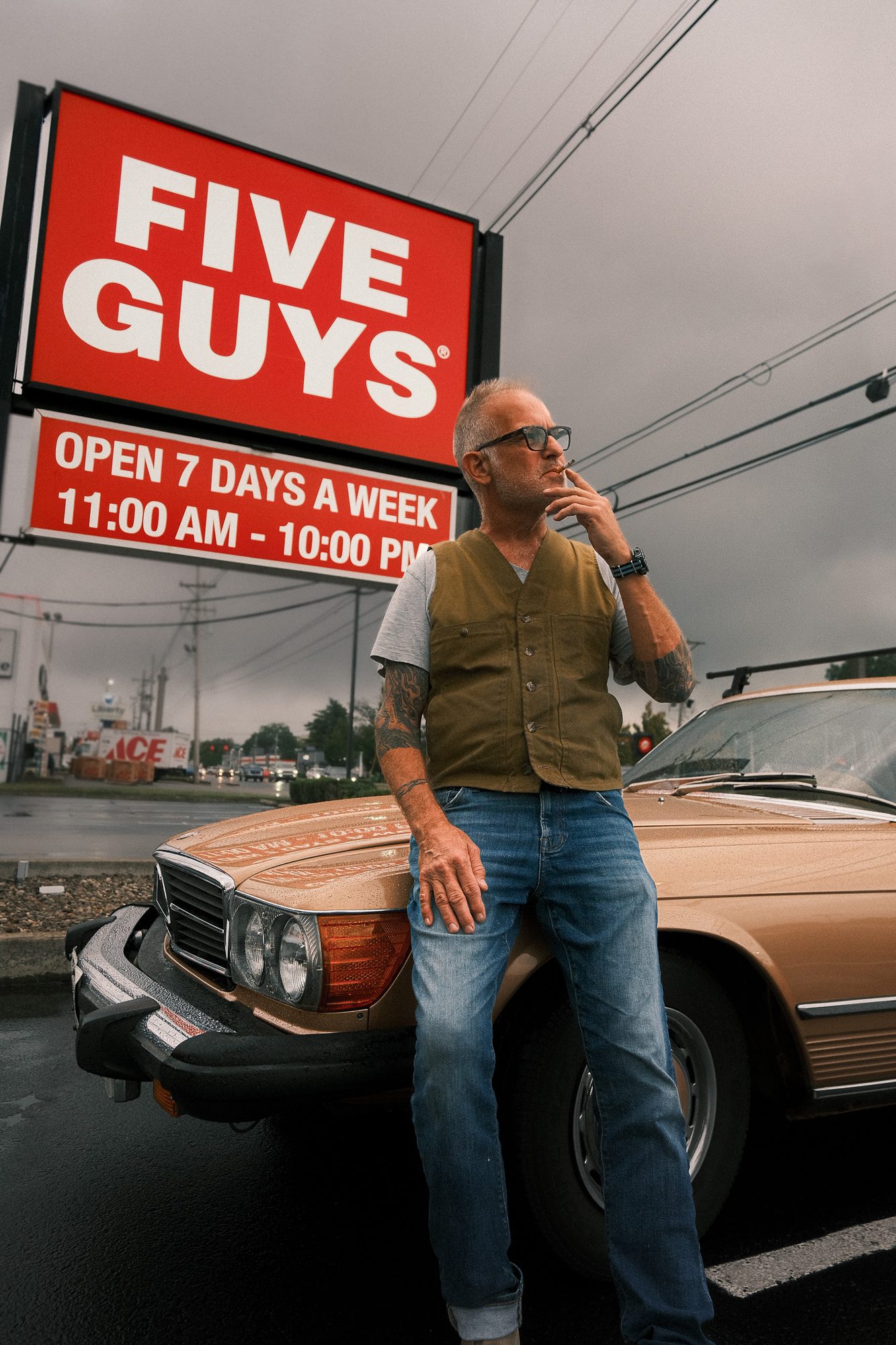 Man in a vest leaning on a vintage Mercedes under a Five Guys sign